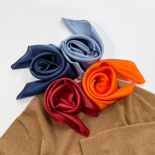 Four silky square scarves in navy blue, sky blue, burgundy, and orange, elegantly arranged on a camel-colored knit garment, showcasing their luxurious sheen.