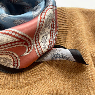 Luxurious silk paisley scarf featuring salmon pink and light blue patterns with intricate designs and black border, draped on camel-colored fabric.