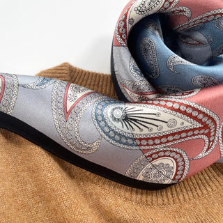Silk bandana scarf with intricate paisley pattern in salmon pink and light blue with black border, displayed on camel-colored knit fabric