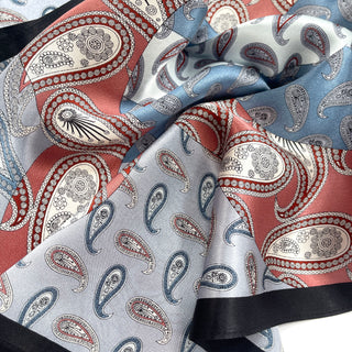 Luxurious silk bandana featuring intricate paisley pattern in salmon pink, light blue, and white with elegant black border, displaying silky texture and folds