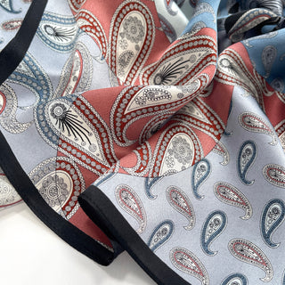Luxurious silk scarf featuring intricate paisley pattern in salmon pink and light blue with elegant black border, showing the fabric's smooth texture and detailed design.
