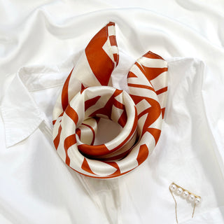 Luxurious burnt orange zigzag silk bandana scarf with creamy white base, elegantly folded on white fabric with pearl pin accessory visible.