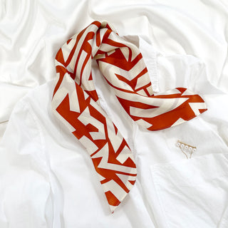 Luxurious burnt orange zigzag pattern silk bandana scarf displayed on white fabric, with a delicate pearl pin accessory nearby