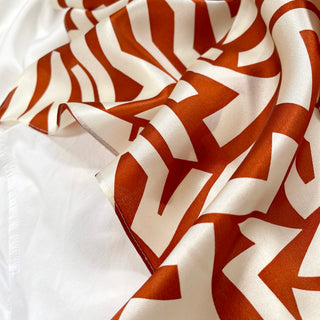 Luxurious silk scarf with burnt orange zigzag pattern on cream white background, showing the fabric's soft drape and glossy texture