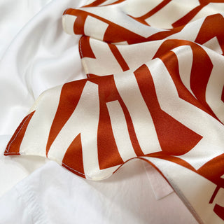 Luxurious silk bandana scarf with burnt orange zigzag pattern on cream white background, showing the fabric's soft texture and lustrous sheen.