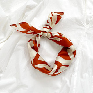 Luxurious silk bandana scarf featuring burnt orange zigzag pattern on cream white background, tied in elegant knot on white fabric