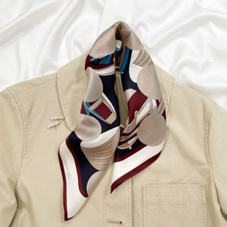 Burgundy Bauhaus silk scarf with geometric patterns in navy, white, and beige, styled as a pocket square on a light beige jacket.
