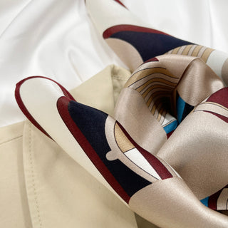 Burgundy Bauhaus Harbor silk scarf with geometric patterns in navy, beige, and turquoise, draped over cream fabric showing its lustrous texture