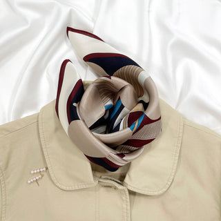 Burgundy Bauhaus Harbor silk scarf styled in a knot on beige coat collar, featuring geometric patterns in navy, burgundy, tan, and blue on luxurious silk fabric