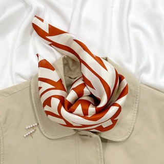 Luxurious silk bandana scarf with burnt orange zigzag pattern on cream white background, styled on beige coat collar with pearl pins