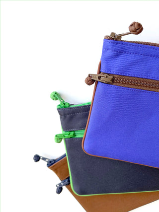 Stacked recycled double zipper wallets in vibrant blue, gray with green trim, and brown colors, displaying neat piping and durable YKK zippers with knotted pulls.