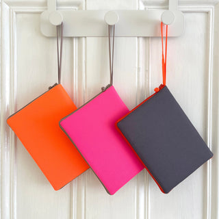 Three recycled Kindle cases hanging on hooks - bright orange, vibrant pink, and dark gray - with wrist straps and zippered closures for travel organization.
