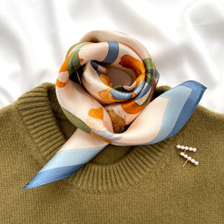 Blue Orange Iris Melody silk scarf elegantly styled on olive green sweater, featuring floral pattern with blue borders and pearl earrings beside it