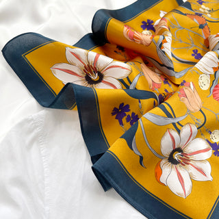 Mustard yellow silk scarf with white flowers, purple accents and butterflies, featuring navy blue border, displayed on white fabric.