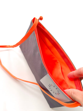 Small gray crossbody wallet with bright orange strap and interior, showing inside pockets and YKK zipper closure, held open to display storage capacity
