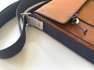 Eco-friendly small messenger bag in orange with navy trim, featuring adjustable strap, brass button closure, and tag showing sustainable materials