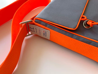 Gray messenger bag with vibrant orange strap and trim detail, featuring a brand tag and adjustable crossbody design on white background.
