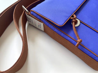 Vibrant blue eco-friendly messenger bag with brown trim and strap, featuring recycled material tag and compact design with decorative cord detail