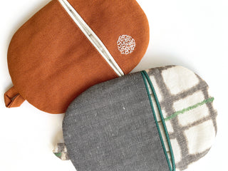 Handmade linen oven mitts set featuring a rust-orange mitt with embroidery detail and a gray herringbone mitt with plaid accent, both with hanging loops