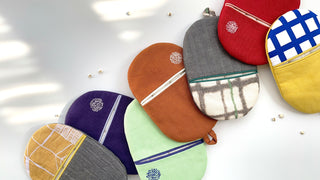Colorful handmade linen oven mitts arranged on white surface, featuring purple, orange, mint, red, and yellow fabrics with decorative embroidery and hanging loops.