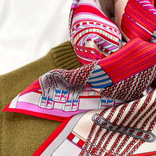 Folded silk scarf with vibrant magenta, red and beige geometric city maze pattern, displaying luxurious texture against olive knit fabric
