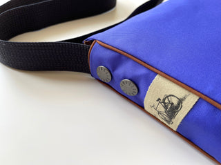 Close-up of vibrant blue recycled crossbody bag with black strap, brown trim, metal studs and fabric label featuring bicycle design