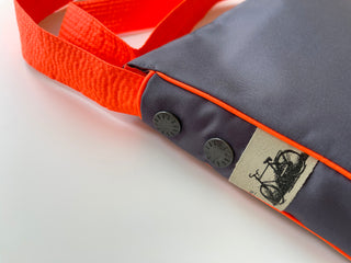 Gray recycled crossbody bag with bright orange strap and trim, featuring decorative metal buttons and bicycle logo tag on fabric patch