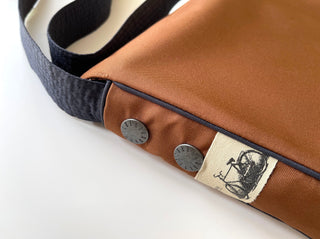 Close-up of recycled crossbody bag in rust orange with navy strap, metal button details, and decorative bicycle logo tag on white background