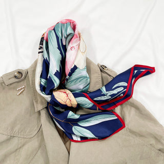 Luxury silk square scarf with navy blue base and vibrant hydrangea floral print in pink, teal and cream, styled on beige trench coat with red border accent