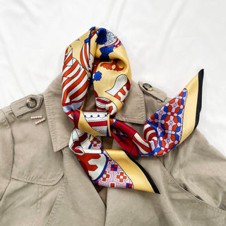 Light yellow silk scarf with medieval carnival pattern in red and blue, elegantly styled on beige trench coat with decorative pin