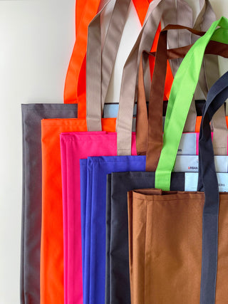 Collection of recycled large tote bags in vibrant colors including orange, pink, blue, green, brown, and gray, with sturdy handles against white background.