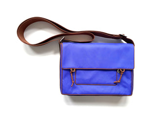 Vibrant blue small messenger bag with brown leather trim and crossbody strap, featuring front flap closure and exterior pocket, set against white background