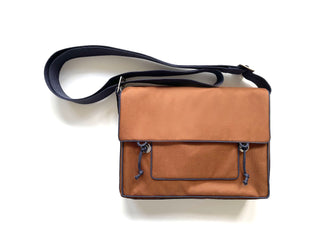 Stylish preppy messenger bag in terracotta brown with navy blue strap and trim, featuring front pocket with drawstring detail, compact rectangular design