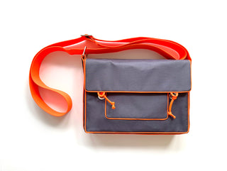 Stylish preppy messenger bag in gray fabric with vibrant orange trim and strap, featuring front pocket with drawstring detail on white background