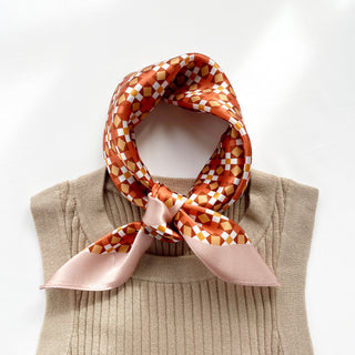 Pink orange silk bandana scarf with retro mosaic cube pattern tied around beige sweater neckline, displaying soft sheen and geometric design
