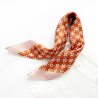 Square silk bandana with orange and pink geometric mosaic pattern, featuring pink border edges, styled in a loose drape on white surface