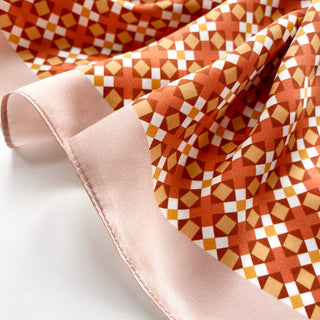 Silk scarf featuring retro mosaic pattern in orange, gold and white with pink border, showcasing the fabric's luxurious sheen and lightweight texture.