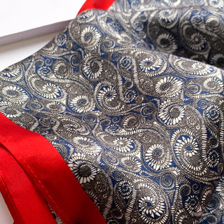 Deep blue silk neckerchief displaying intricate spiral wave pattern in white and gray, with vibrant red border accent showing luxurious fabric texture