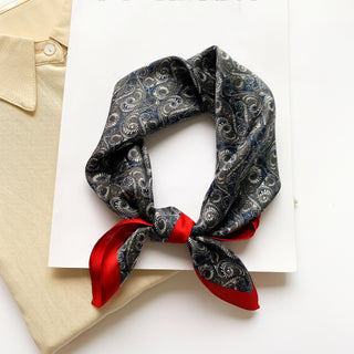 Deep blue silk neckerchief with spiral wave pattern, vibrant red border, elegantly tied and displayed on white background beside beige fabric