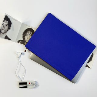 Vibrant blue recycled laptop sleeve covering MacBook with zippered closure, shown with earbuds and magazine on white background