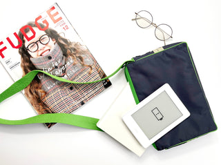 Navy blue recycled crossbody bag with bright green strap and trim, displayed with e-reader, glasses, and magazine on white background