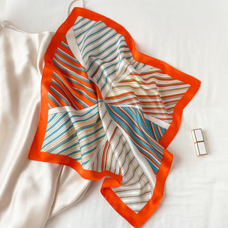 Square silk scarf with bright orange border and sunburst striped pattern in navy blue, orange and cream, elegantly displayed on white fabric