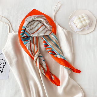 Silk bandana scarf with vibrant orange border and blue, beige, and white striped pattern, elegantly folded on cream fabric
