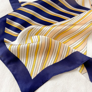 Luxurious silk bandana with navy border and striped pattern in orange, yellow and cream, displaying soft folds that highlight its silky texture