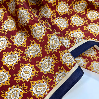Luxurious silk neckerchief featuring rich red background with gold and white paisley pattern, bordered with navy blue trim showing elegant fabric texture.