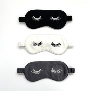 Three luxury silk sleep masks featuring embroidered eyelash designs - black, white, and dark gray - with adjustable elastic bands for comfort