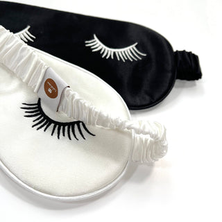 Luxury double-sided silk sleep mask featuring black and white sides with embroidered eyelash design, elastic band, and quality tag visible