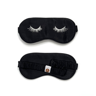 Luxurious black silk sleep mask with white embroidered eyelashes, shown from front and back views with elastic band and brand tag.