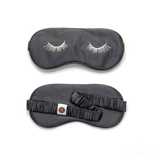 Black silk sleep mask with white embroidered eyelashes shown from front and back, featuring elastic band with brand tag for comfortable fit