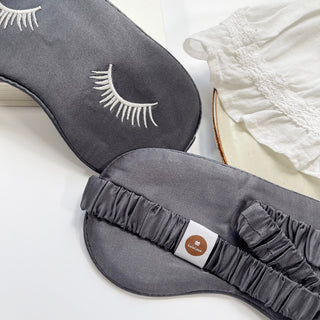 Double-sided silk sleep mask in dark gray with white embroidered eyelash design, showing elastic band with brand tag, beside white fabric.